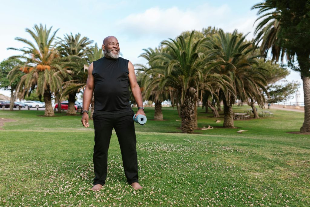 Elderly African American man doing yoga with a mat in a sunny park.